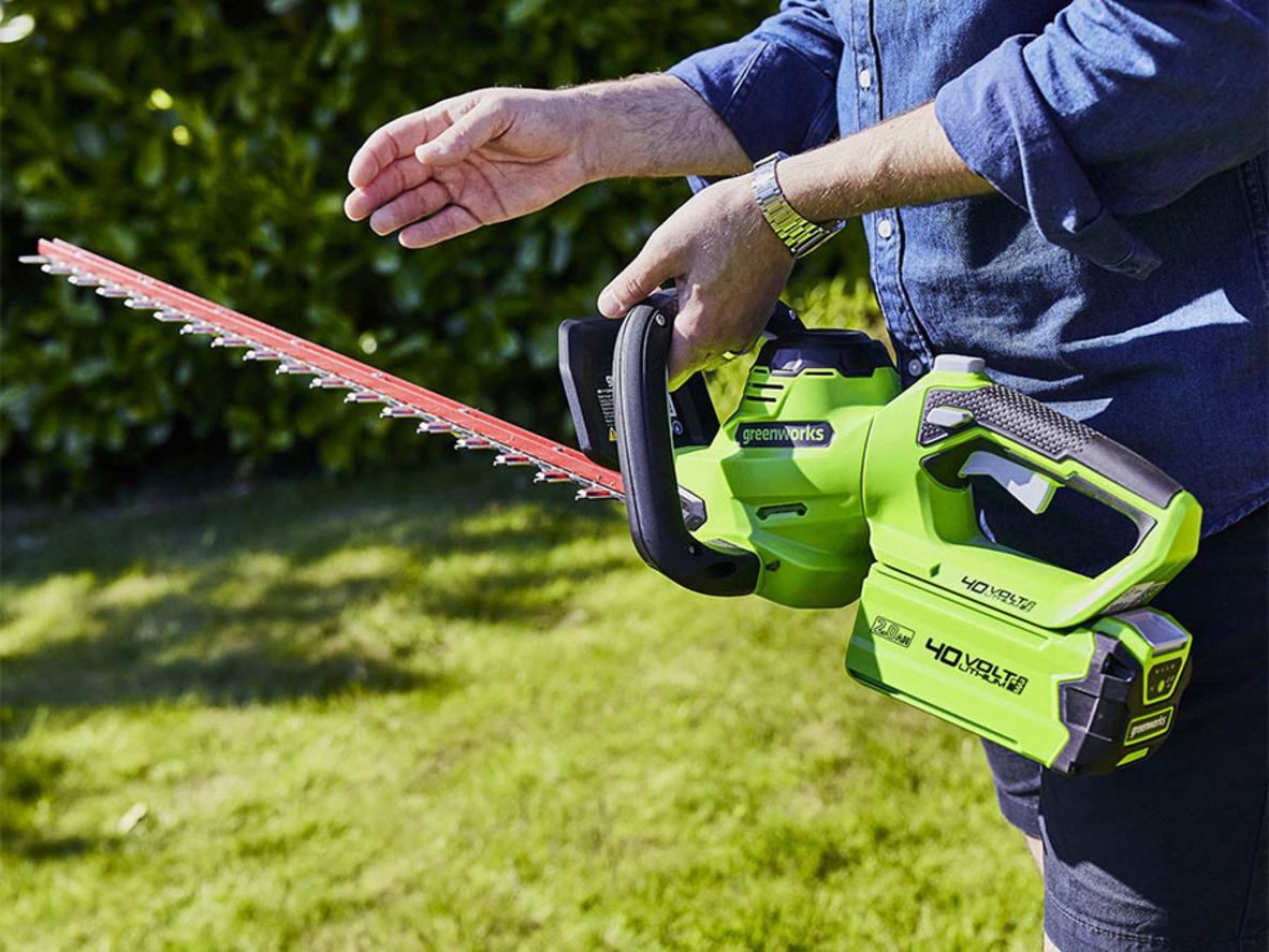 Người dùng thao tác trên thang để tỉa phần hàng rào cao, imagePrompt: User trimming a hedge overhead with the rotating handle set for vertical cutting.